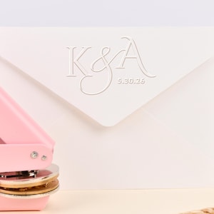 Puede incluir: Sobre de color marfil con las iniciales "K&A" en relieve y la fecha "5.30.25". Un sello de sobre rosa y un vaso decorativo están en el fondo. Una espiga de trigo está en primer plano. El sobre es para invitaciones de boda.