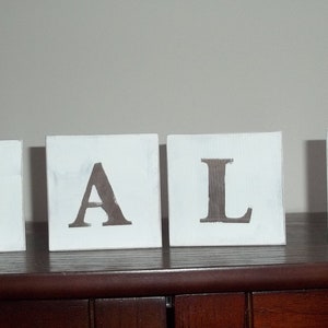Puede incluir: Cuatro bloques cuadrados blancos que deletrean la palabra "FALL" en letras marrones. Los bloques están dispuestos uno al lado del otro sobre una superficie de madera oscura, con una pared neutra en el fondo.