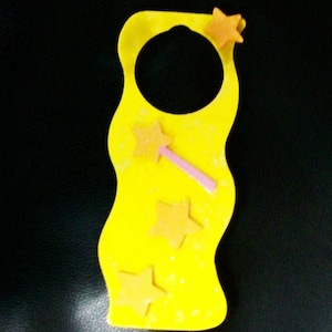 May include: A yellow, wavy-edged craft with a large central hole and three orange star accents. A pink wand with a star is also attached. The craft is decorated with small white speckles and is set against a black background.