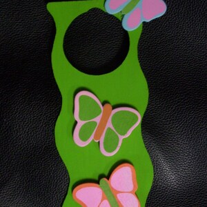 Peut inclure: Un accroche-porte vert avec un design ondulé et une ouverture circulaire. Il présente trois papillons colorés roses, verts et oranges. Les papillons sont de tailles et de positions variées, ajoutant une touche fantaisiste.