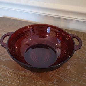 May include: A ruby red glass bowl with two ornate handles. The bowl has a circular shape with a ribbed exterior and a starburst pattern on the base. This vintage bowl is perfect for serving salads or fruit.