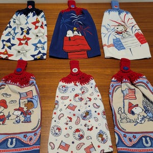 May include: Six hanging kitchen towels featuring Peanuts characters and patriotic themes. Each towel has a crocheted top in red, white, or blue with a matching button. The fabric prints include Snoopy, Woodstock, American flags, stars, and fireworks.