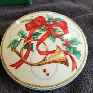 May include: A round, cream-colored decorative box lid featuring a red bow, holly leaves, and a golden horn. The design includes red berries and a gold rope border, evoking a festive holiday theme.