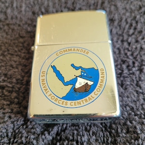 May include: A silver-colored lighter with a hinged lid. The lighter features a circular emblem with the text "COMMANDER" and "US NAVAL FORCES CENTRAL COMMAND" around a blue and brown nautical design.