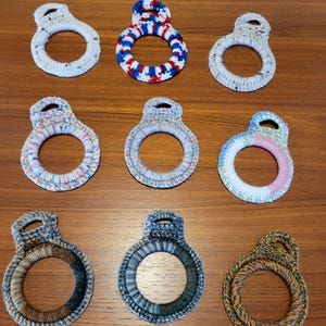 May include: Nine handmade crochet towel rings in various colors and patterns. The rings feature a circular design with a loop at the top for hanging. Colors include white, blue, red, pink, and brown. Each ring is unique, showcasing the artistry of handmade crafts.