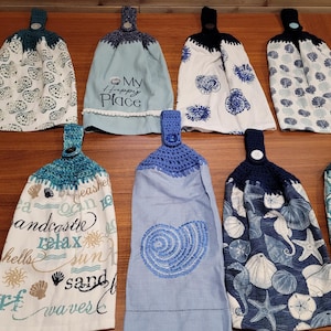 May include: A collection of hanging kitchen towels with crochet tops. The towels feature various sea-themed patterns, including seashells, seahorses, and starfish, in shades of blue, white, and beige. Some towels have embroidered text, such as "My Happy Place."