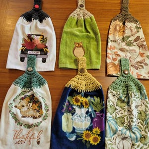 May include: Six decorative kitchen hand towels with crocheted tops. The towels feature various autumn-themed designs, including a truck with flowers, an owl, sunflowers, a hedgehog, and pumpkins. Each towel has a button closure.