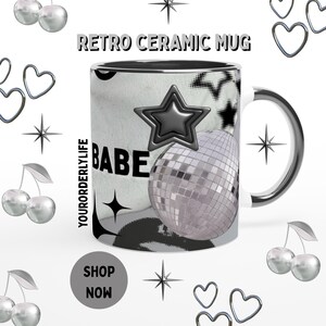 Könnte beinhalten: Retro-Keramikbecher mit silberfarbenem Henkel und Rand. Der Becher hat ein Schwarz-Weiß-Design mit einem Stern, einer Discokugel und dem Wort "BABE". Der Text "RETRO CERAMIC MUG" steht oben und "YOURORDERLYLIFE" an der Seite.