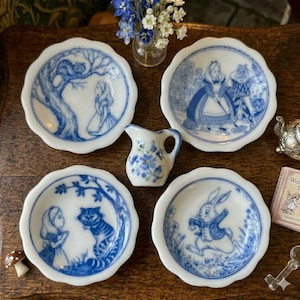 May include: A collection of miniature white and blue porcelain plates and a small pitcher. Each plate features a different scene from Alice in Wonderland. The plates have a scalloped edge. The pitcher has a floral design.