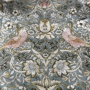 May include: A fabric textile featuring a repeating pattern of two birds facing each other, surrounded by intricate floral and leaf motifs in shades of muted blue, cream, soft pink, and olive green. The design includes symmetrical blossoms and foliage.