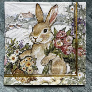 May include: A decorative paper napkin featuring a watercolor illustration of a brown rabbit holding a basket of flowers. The rabbit is set against a backdrop of a white fence, a house, and birds. The napkin has a gold border.