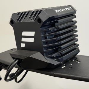 Può includere: Un dispositivo elettronico nero con la scritta "FANATEC" stampata in alto. Il dispositivo ha una serie di creste orizzontali e un logo con linee bianche. I cavi si estendono dalla parte inferiore del dispositivo, montato su una superficie metallica nera.