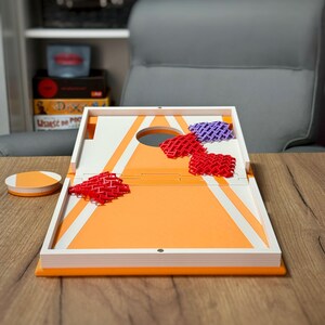 May include: A tabletop cornhole game set featuring a white and orange board with a circular hole. The set includes a small orange and white circular disc and several flexible, lattice-patterned bean bags in red and purple colours.