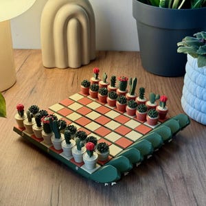 Può includere: Un originale set di scacchi a tema cactus. La scacchiera presenta un motivo a scacchi rosso e crema. I pezzi sono cactus in miniatura in piccoli vasi bianchi e terracotta. La scacchiera ha un bordo verde e i pezzi sono disposti per giocare.