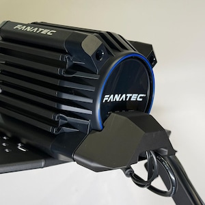 Könnte beinhalten: Schwarze Fanatec-Rennradkomponente mit dem Markennamen in Weiß. Die Komponente hat ein geripptes Design und einen blauen Akzentring. Sie ist auf einer schwarzen Plattform montiert.