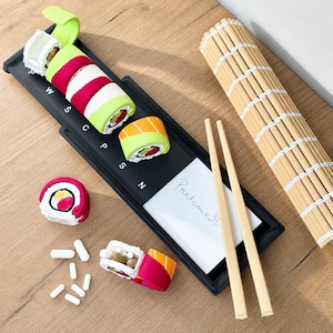 Könnte beinhalten: Ein schwarzes rechteckiges Tablett mit den aufgedruckten Buchstaben W, S, C, P, S, N, das mehrere sushi-förmige Gegenstände in Rot, Grün, Weiß und Orange enthält. Eine weiße Karte mit dem Text Printomix 3D, zwei Holzstäbchen und eine Bambusmatte.