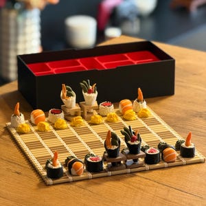 Könnte beinhalten: Ein Schachspiel im Sushi-Design mit Spielfiguren in Form verschiedener Sushi-Rollen, Nigiri und Handrollen, angeordnet auf einem Bambusmatten-Spielbrett. Das Set enthält eine schwarze Aufbewahrungsbox mit rotem Inneneinsatz für die Miniaturteile.