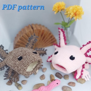 Può includere: Due peluche di axolotl all'uncinetto, uno marrone e uno rosa, con un verme rosa all'uncinetto davanti a loro. Gli axolotl hanno occhi neri e bocca aperta. I peluche sono seduti su una superficie bianca con piccoli sassi intorno. L'immagine include il testo "PDF pattern" e "NoodleLoops".
