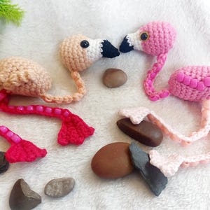 May include: Crocheted flamingo ornaments in shades of pink and tan, with black and white beaks. The flamingo's legs are adorned with pink beads. The ornaments are placed on a white surface with decorative stones.