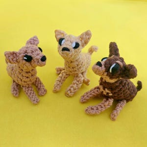 May include: Three handmade crochet chihuahua dog figurines in shades of brown and tan, with black eyes and noses, sit on a yellow background. The dogs are in a playful pose, showcasing their unique crochet texture.