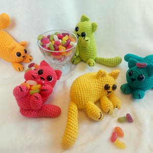 May include: Five colorful crochet cat toys, each with large black eyes and small paws. One orange cat stretches out, while a pink cat holds jelly beans. A green, yellow, and teal cat are also present, with a glass bowl of jelly beans in the background.
