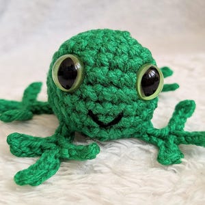 May include: A handmade green crochet frog with large, round, black-pupiled eyes and a smiling mouth. The frog has four small, webbed feet and is made of a textured yarn. The frog is a whimsical, handcrafted item.
