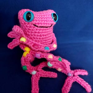 May include: A pink crocheted frog toy with blue eyes and colourful beads on its legs.