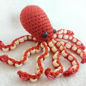May include: A handmade, crocheted octopus in shades of red. The octopus has eight tentacles, each adorned with white beads and pink accents. The eyes are small and black. This soft toy is likely a children's item.