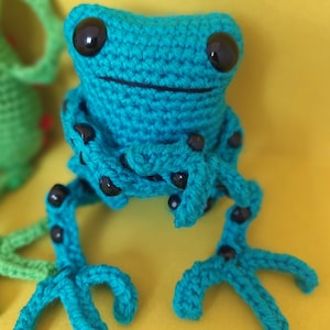May include: A crocheted green frog with black eyes and black accents on its limbs. The frog is sitting on a yellow surface.