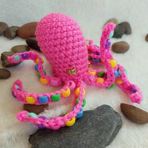 Puede incluir: Un pulpo rosa hecho a mano con ganchillo y tentáculos con cuentas de varios colores. El pulpo está sentado sobre una roca gris oscura, con otras rocas y un fondo blanco y esponjoso.