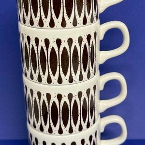 May include: A stack of four white ceramic mugs with a brown, teardrop-shaped pattern. Each mug has a curved handle and a simple, clean design. The mugs are stacked on top of each other against a blue background.