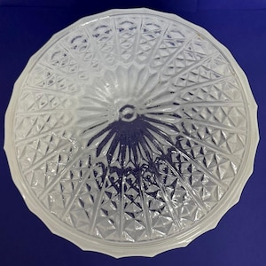 May include: A clear, round glass ceiling light cover with a detailed geometric pattern. The cover has a central hole and a scalloped edge. The design features diamond and triangular shapes radiating from the center.
