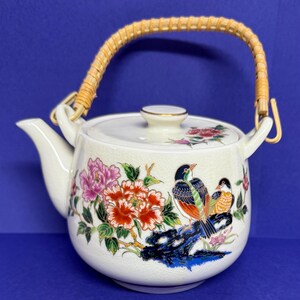 May include: Cream ceramic teapot with a woven handle. The teapot is decorated with colourful floral and bird designs. The handle is made of woven straw. The teapot has a spout and a lid with a gold-coloured trim.