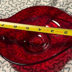Puede incluir: Un cuenco de vidrio rojo vibrante con una forma única de hoja. El cuenco mide aproximadamente 20 cm de largo y presenta un patrón texturizado. El interior tiene una hendidura circular. Una cinta métrica amarilla está colocada sobre el cuenco.