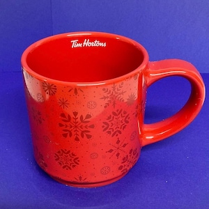 May include: A red ceramic mug with a handle, decorated with a snowflake pattern and the "Tim Hortons" logo. The mug is a bright red colour and is designed for hot drinks.