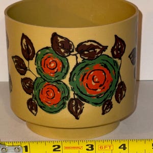 May include: A yellow plastic container with a floral design. The flowers are red and green with brown leaves. The container is approximately 10 cm tall.