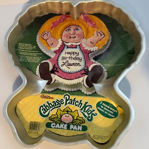 Könnte beinhalten: Eine Vintage Cabbage Patch Kids Kuchenform. Die Form hat die Form eines Cabbage Patch Kids mit einem rosa Kleid und den Worten "Happy Birthday Lauren". Die Verpackung trägt die Aufschrift "Cake Pan" und "5 more ways to decorate shown on back!"
