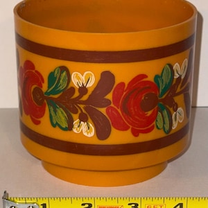 May include: Orange plastic container with a floral design. The container has a cylindrical shape with a base. The floral design includes red roses, green leaves, and white accents. Brown bands encircle the top and bottom.