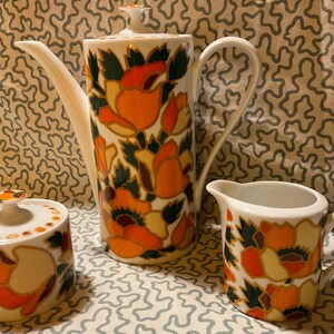 May include: A vintage ceramic coffee set comprising a coffee pot, creamer jug, and sugar bowl. The set is adorned with an orange, yellow, and green floral design on a white base. The coffee pot has a long spout and a lid.
