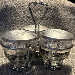 Peut inclure: Un ensemble à condiments argenté avec trois bols en verre transparent. Chaque bol a un couvercle argenté décoratif avec un motif de rose et une bande à motifs. Une poignée en forme de cœur relie les bols.