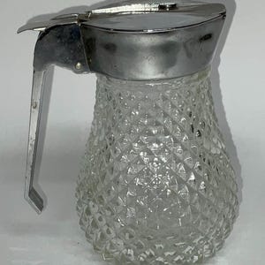 May include: A clear glass syrup jug with a diamond pattern and a silver-coloured metal lid and handle. The jug has a spout for easy pouring and is designed for serving syrup or other condiments.