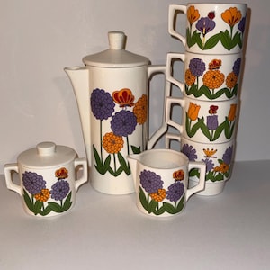 May include: A vintage white ceramic tea set with a floral design. The set includes a teapot, sugar bowl, creamer, and six cups. The flowers are purple, orange, and yellow with green leaves. The set is likely from the 1970s.