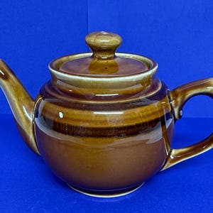 May include: Brown ceramic teapot with a lid and handle. The teapot has a rounded body and a spout. The color is a gradient of brown hues. The teapot is set against a blue background.