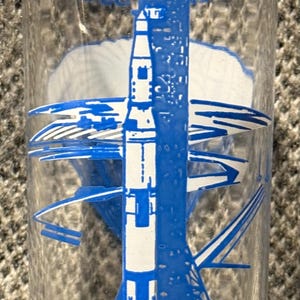 Könnte beinhalten: Klares Glas mit einer blau-weißen Grafik einer startenden Rakete. Die Worte "BLAST-OFF" sind unten aufgedruckt. Das Design umfasst die Rakete, den Startturm und stilisierte Abgase.