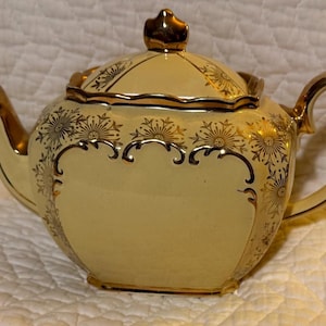 Könnte beinhalten: Cremefarbene Teekanne mit goldenen Akzenten. Die Teekanne hat einen runden Körper, einen gebogenen Griff und einen Ausguss. Goldene Blumenmuster und Verzierungen zieren Deckel und Körper. Wahrscheinlich aus Keramik oder Porzellan.