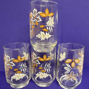 May include: Four clear drinking glasses with a leaf pattern. The leaves are white, gold, and brown, with fine vein details. The glasses are cylindrical with a slightly flared base, suitable for serving drinks.