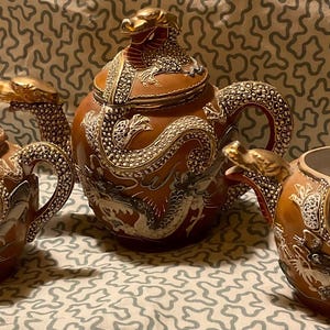 Op de afbeelding: Een driedelige keramische theeset met een drakenmotief. De set bevat een theepot met deksel en twee kleinere theekopjes. De stukken zijn bruin met witte en gouden draken details. De handvatten en tuiten zijn gevormd als drakenkoppen.