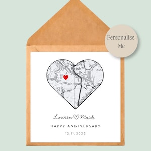 May include: A square white card inside a brown kraft paper envelope featuring a heart-shaped map with a red heart icon. The text reads Lauren, a heart symbol, Mark, HAPPY ANNIVERSARY, and 12.11.2022. A circular beige sticker says Personalise Me.