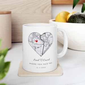 Op de afbeelding: Een witte keramische mok met een hartvormige kaartafbeelding en een rood hartpictogram. Onder de kaart staat de tekst Paul ♡ David, WHERE YOU SAID YES, en de datum 12.11.2022 in zwarte letters. De mok is voorzien van een handvat aan de zijkant.