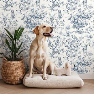 May include: A golden Labrador sits on a beige dog bed in front of a wall with a blue and white dog-themed toile pattern. A woven basket with a green plant sits to the left. A stuffed dog toy rests on the bed.